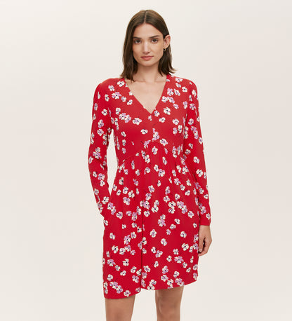 A woman in the Joanne Red Poppies Jersey Short Dress, featuring long sleeves, a V-neckline, and a white floral print, stands against a plain light background, embodying effortless everyday comfort.