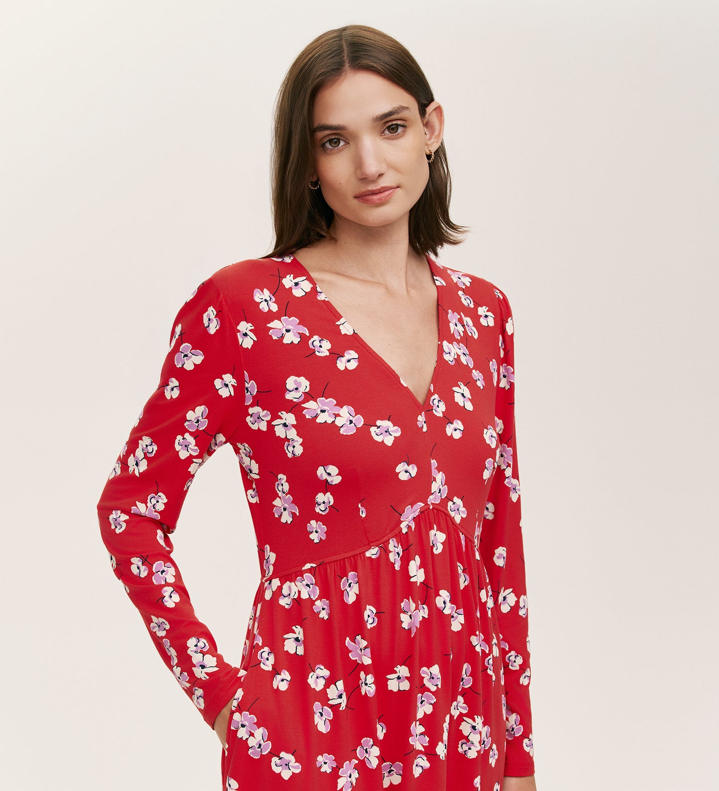 A woman with straight brown hair confidently models the Joanne Red Poppies Jersey Short Dress, featuring long sleeves and a red floral print, standing against a plain light background and showcasing comfortable, everyday style.