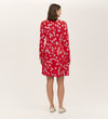 A woman faces away, wearing the Joanne Red Poppies Jersey Short Dress with white floral print and beige flats, ideal for everyday comfort. The background is plain and light-colored.