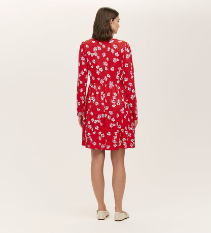 A woman faces away, wearing the Joanne Red Poppies Jersey Short Dress with white floral print and beige flats, ideal for everyday comfort. The background is plain and light-colored.
