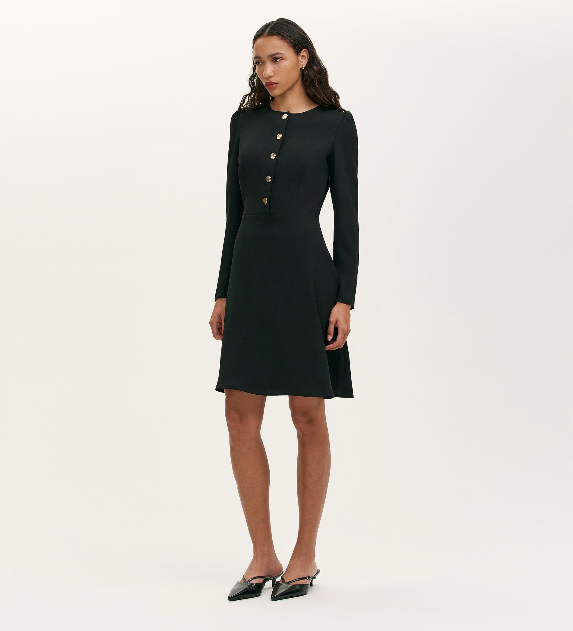 A woman wears the Kadie Black Crepe Short Dress, featuring long sleeves, knee-length cut, and gold button details, while standing against a white background. She has long wavy hair and pairs the dress with black pointed-toe shoes.