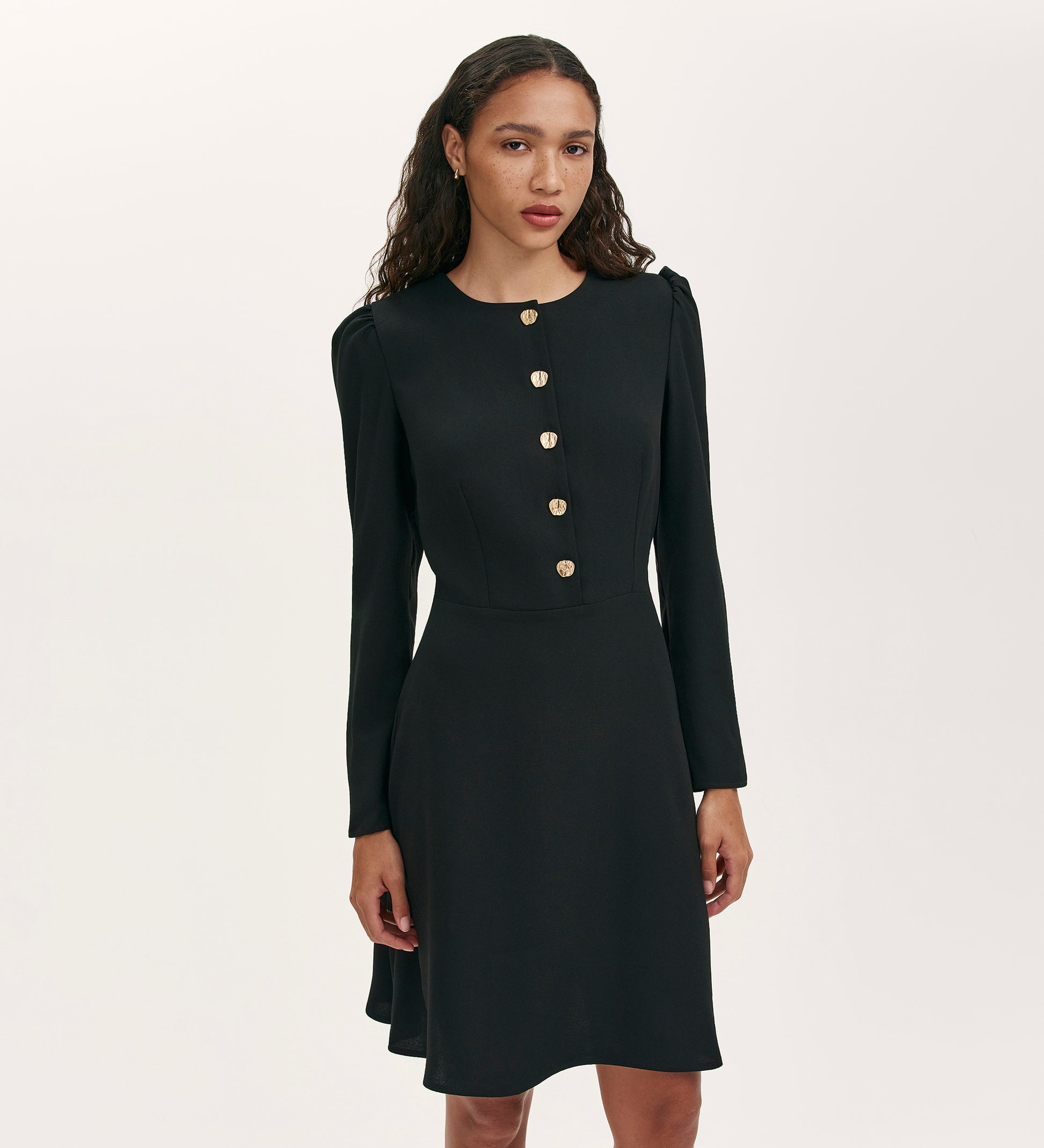A woman with long, wavy hair wears the Kadie Black Crepe Short Dress with gold buttons down the front, standing against a plain light background.