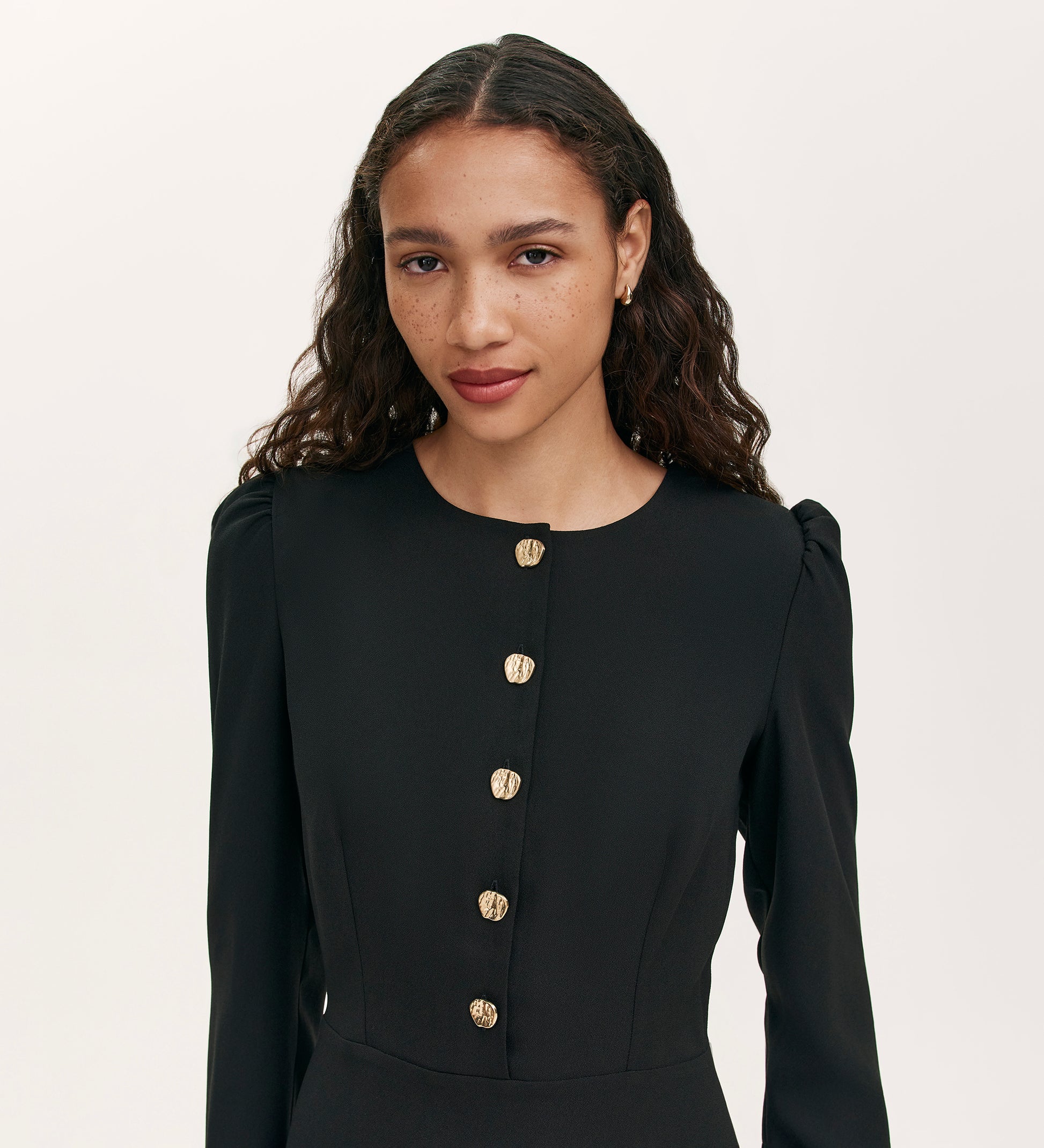A woman with long, wavy brown hair wears the Kadie Black Crepe Short Dress featuring gold button details, standing against a plain light background. She has a calm expression and gazes slightly to the side.