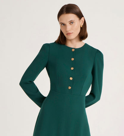 A woman with straight brown hair wears the Kadie Bottle Green Crepe Short Dress, which has long sleeves and gold buttons down the front. She stands against a plain light background, looking directly at the camera.