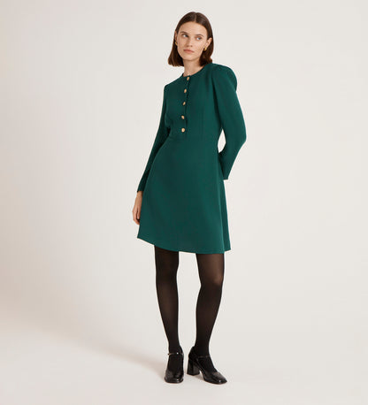A woman with straight brown hair and a neutral expression stands against a plain background wearing the Kadie Bottle Green Crepe Short Dress with gold button details, black tights, and black shoes.