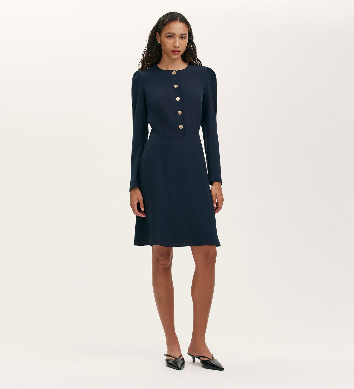 A woman faces forward in the Kadie Navy Crepe Short Dress, featuring long sleeves, a fitted waist, gold front buttons, and black pointed-toe heels, set against a plain light background.