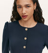 A woman with long wavy hair and freckles wears the Kadie Navy Crepe Short Dress, featuring gold button details and puffed shoulders, looking calmly past the camera.