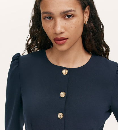 A woman with long wavy hair and freckles wears the Kadie Navy Crepe Short Dress, featuring gold button details and puffed shoulders, looking calmly past the camera.