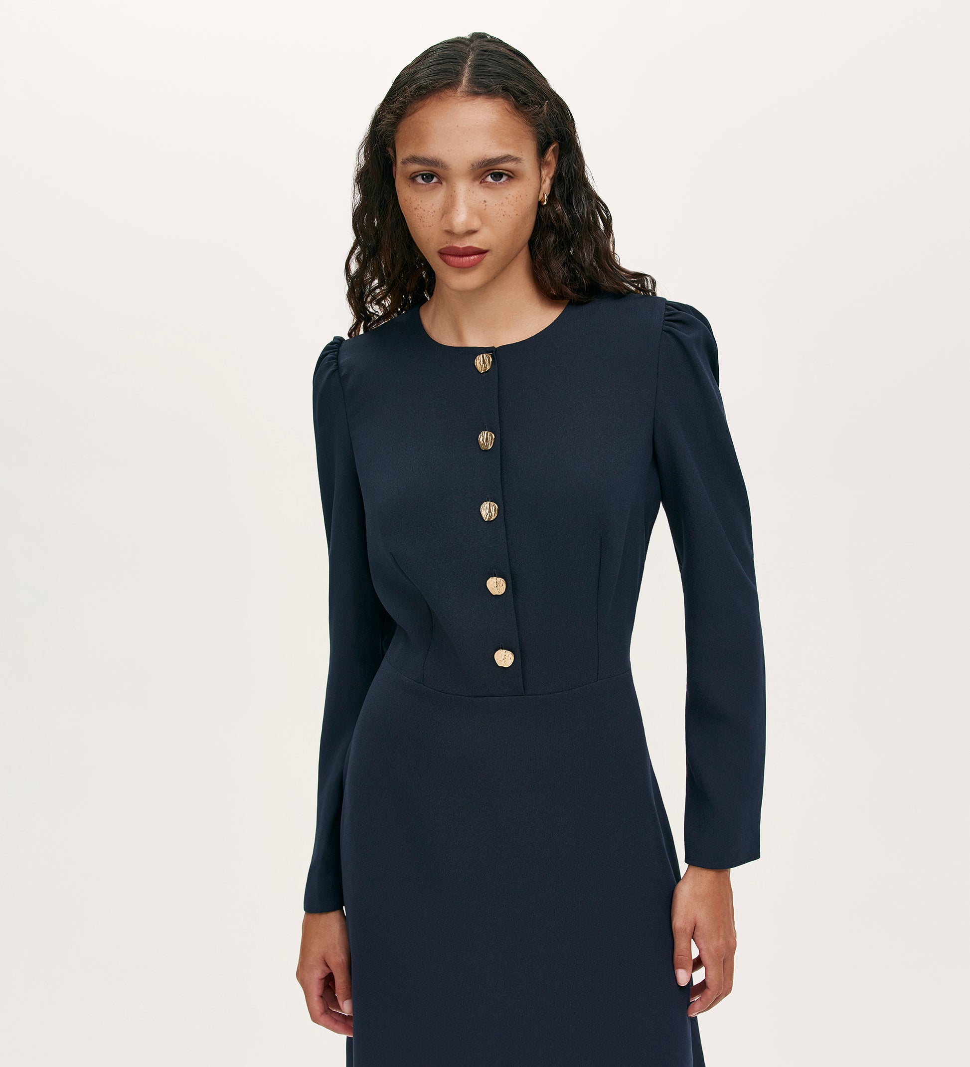 A woman with long, wavy hair models the Kadie Navy Crepe Short Dress, a fitted mini dress with gold button detailing, against a plain light background.