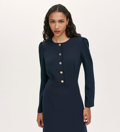 A woman with long, wavy hair models the Kadie Navy Crepe Short Dress, a fitted mini dress with gold button detailing, against a plain light background.