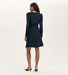 A woman faces away, wearing the Kadie Navy Crepe Short Dress—knee-length with long sleeves, slight shoulder padding—and black low-heeled shoes. The background is plain and light-colored.