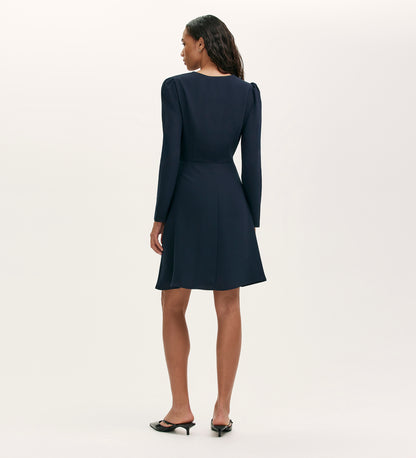 A woman faces away, wearing the Kadie Navy Crepe Short Dress—knee-length with long sleeves, slight shoulder padding—and black low-heeled shoes. The background is plain and light-colored.