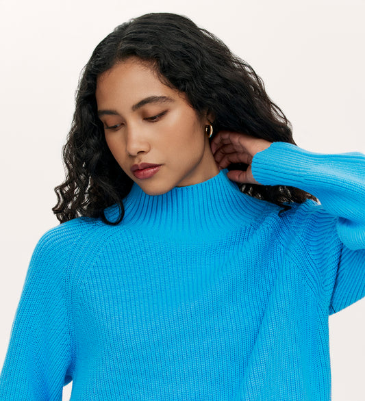 A person with long, curly black hair wears the Kaela Light Blue High Neck Jumper, looking down thoughtfully with one hand touching their neck against a plain light background.