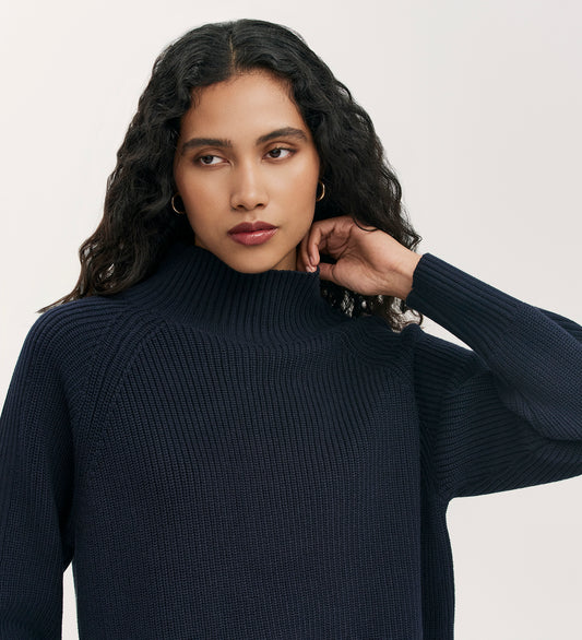 A woman with long, wavy dark hair poses against a light background, wearing the Kaela Navy High Neck Jumper and gold hoop earrings—a timeless women’s wardrobe staple.
