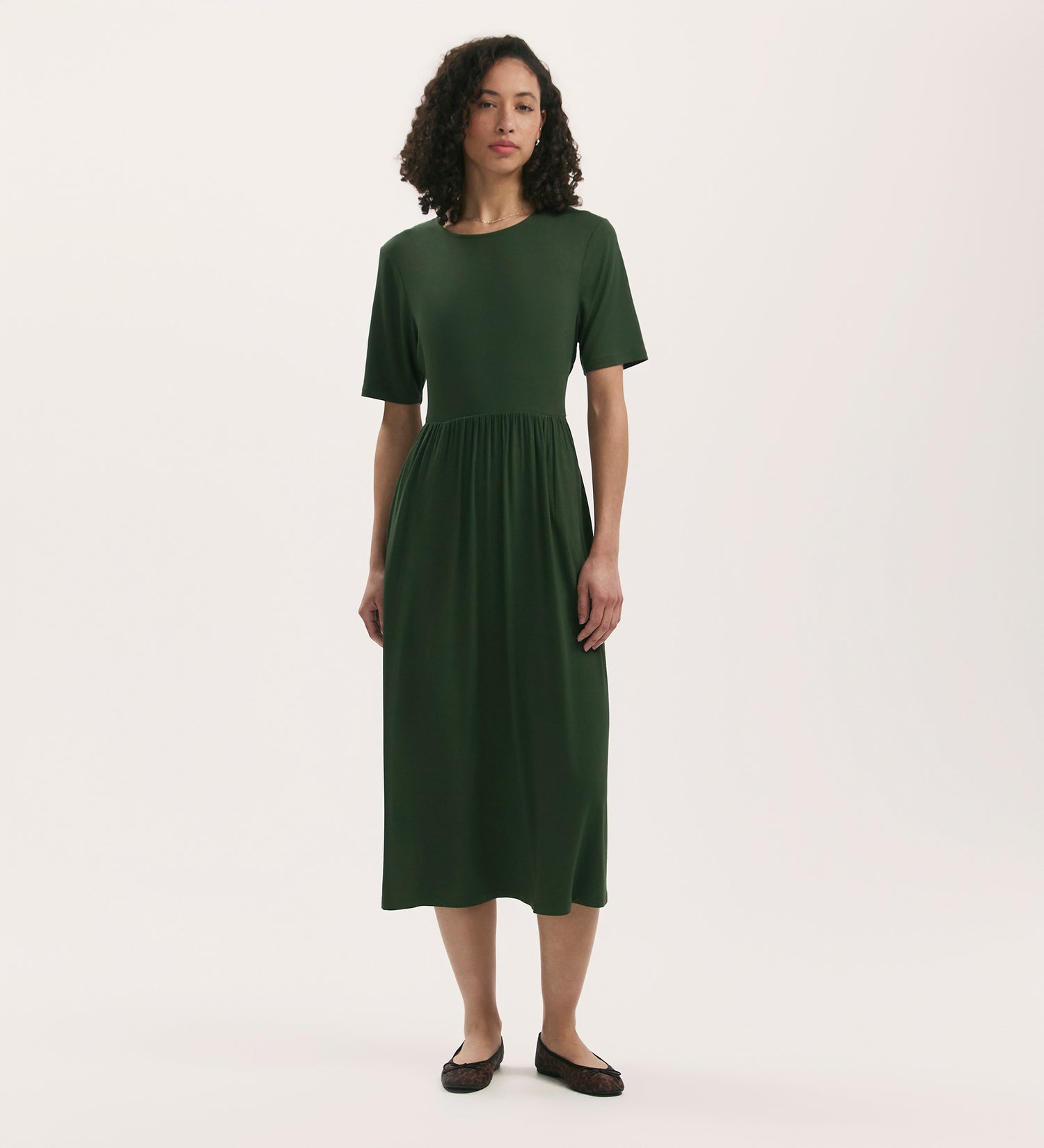 A woman with curly hair wears the Kathryn Khaki Jersey Midi T-Shirt Dress with a relaxed fit and gathered waist, paired with black flats, standing against a plain white background.