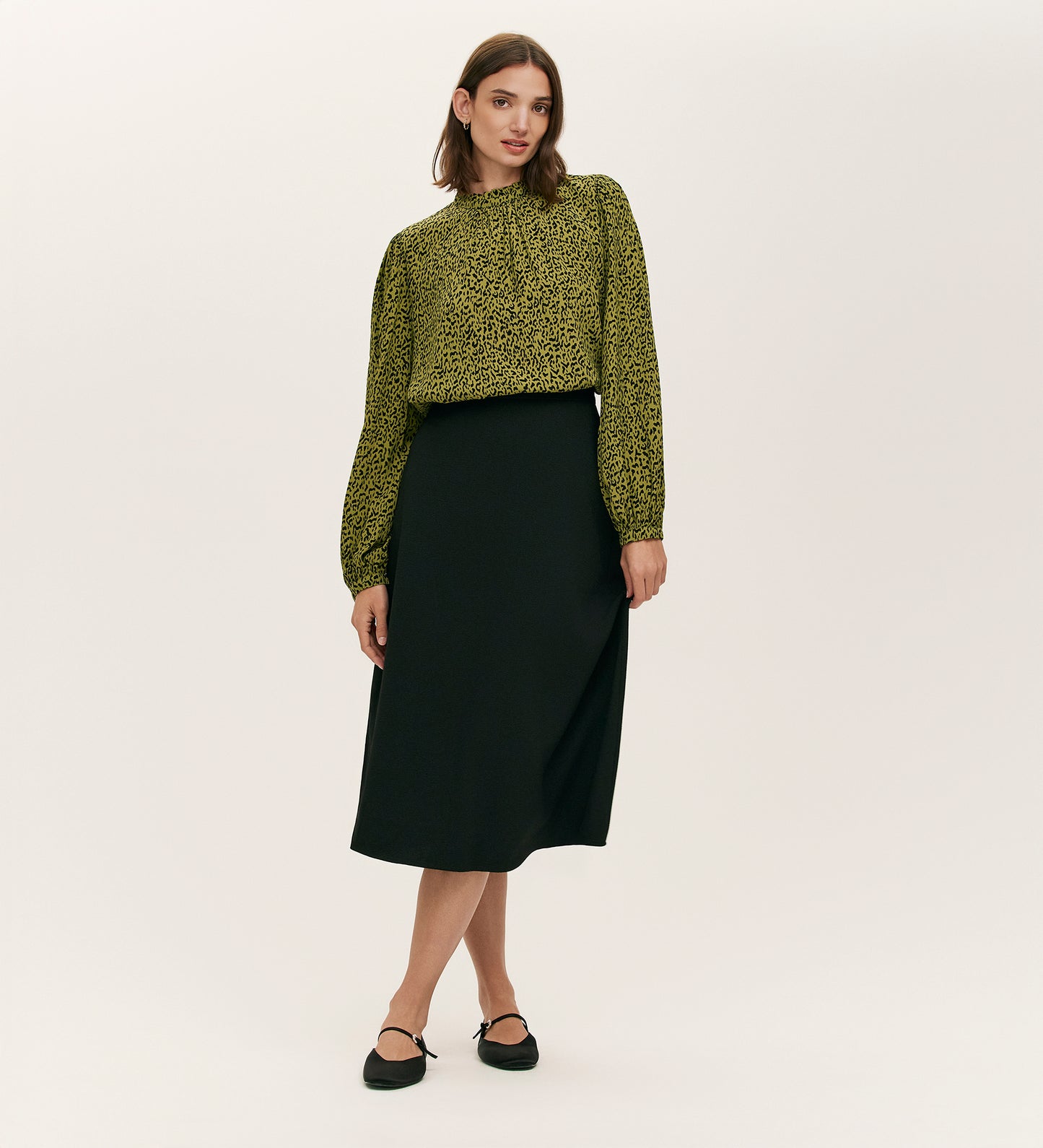 A woman stands against a plain background wearing the Keira Green Leopard Crepe Top, tucked into a high-waisted black midi skirt. She completes the look with black ankle-strap flats—a stylish, romantic wardrobe staple.
