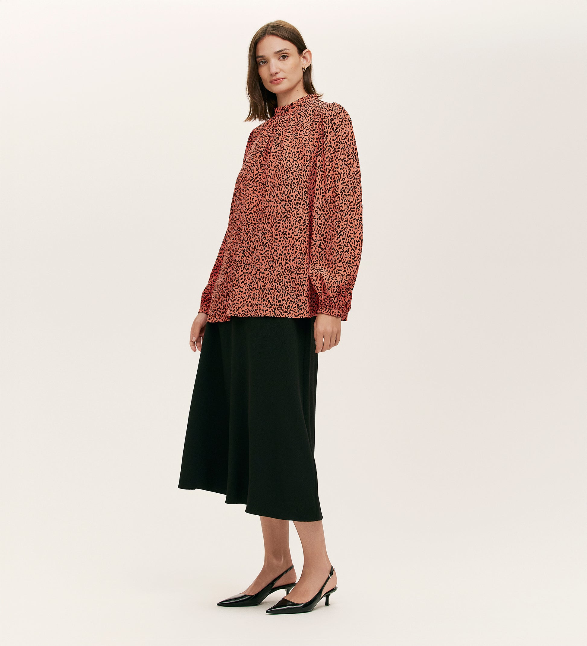 A woman stands against a plain background wearing the Keira Orange Leopard Crepe Top, paired with a black midi skirt and black low-heeled slingback shoes.