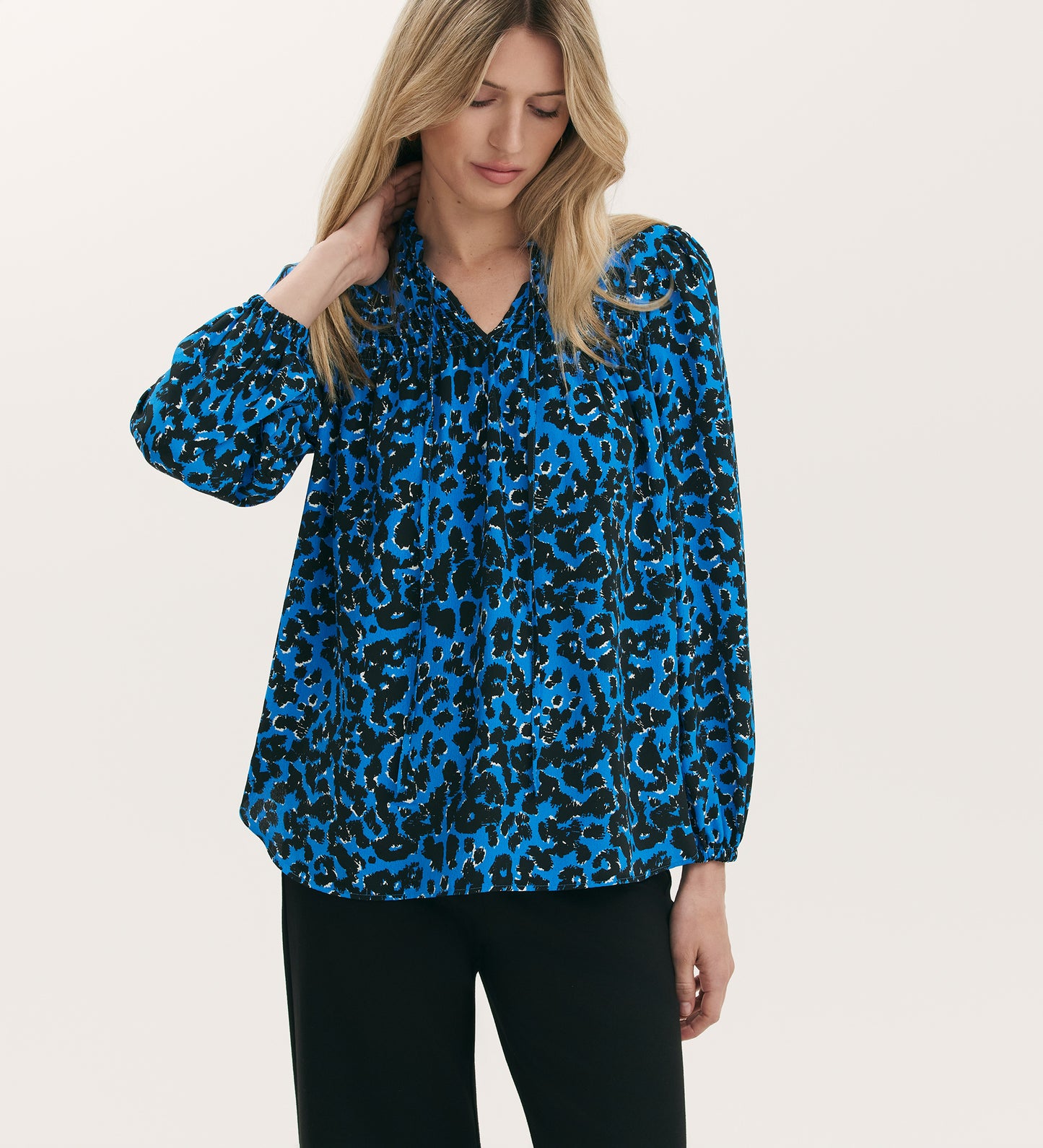 A woman in a Kiya Blue Animal Crepe Long Sleeve Blouse with neckline frills and black pants stands against a plain light background, looking down and touching her hair. The blouse’s relaxed fit adds effortless style to her look.