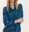 A woman with long blonde hair wears the Kiya Blue Animal Crepe Long Sleeve Blouse, featuring neckline frills and a relaxed fit, standing with her arms crossed against a plain light background.