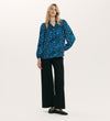 A woman stands against a plain background in the Kiya Blue Animal Crepe Long Sleeve Blouse with neckline frills, paired with black wide-leg pants and studded flats. Her long blonde hair and relaxed fit create a laid-back yet stylish look.