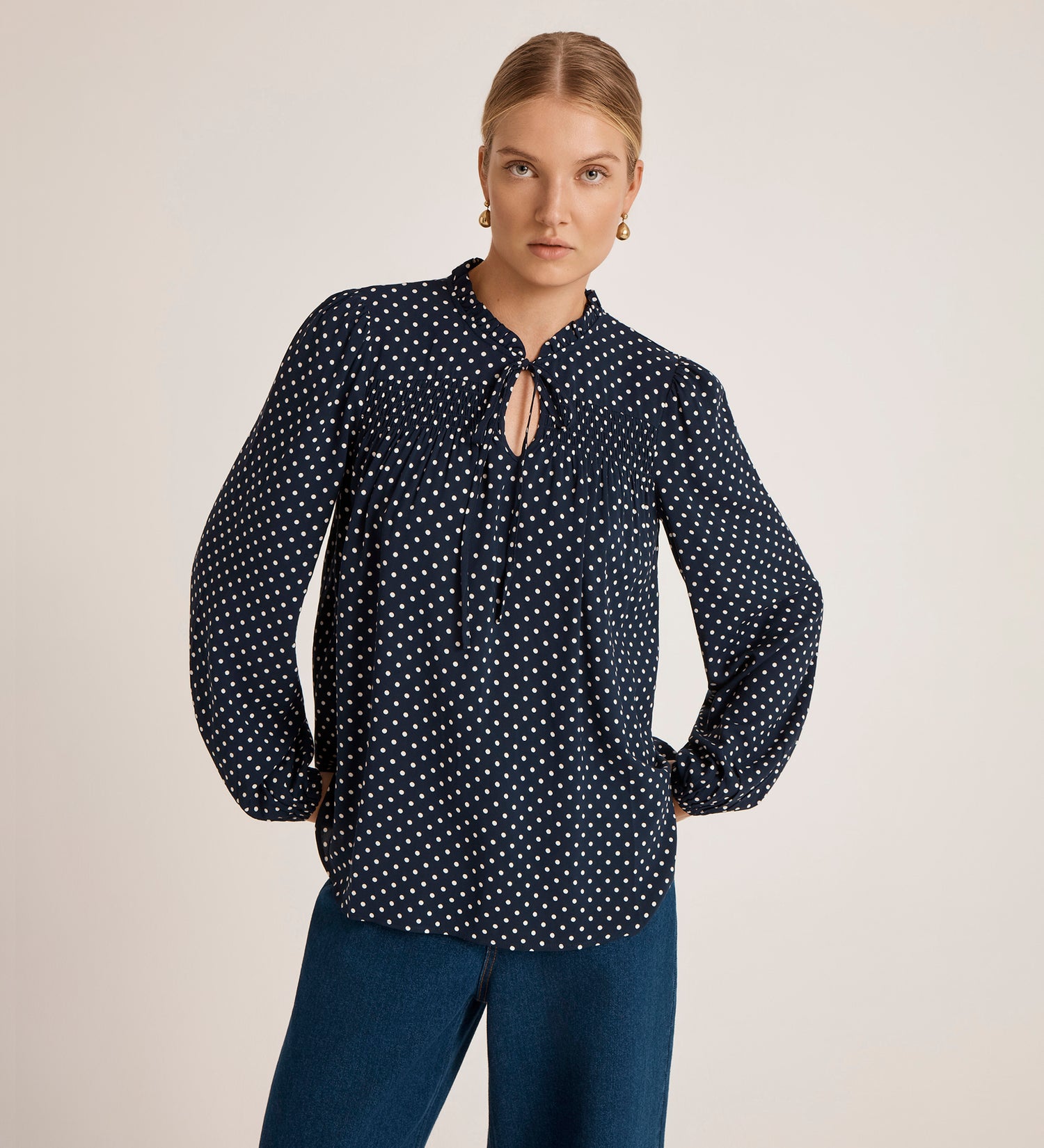 A person wears the Kiya Navy Polka Dot Blouse—long-sleeve, crepe fabric with white polka dots and a keyhole neckline—paired with blue jeans, standing against a plain light background.