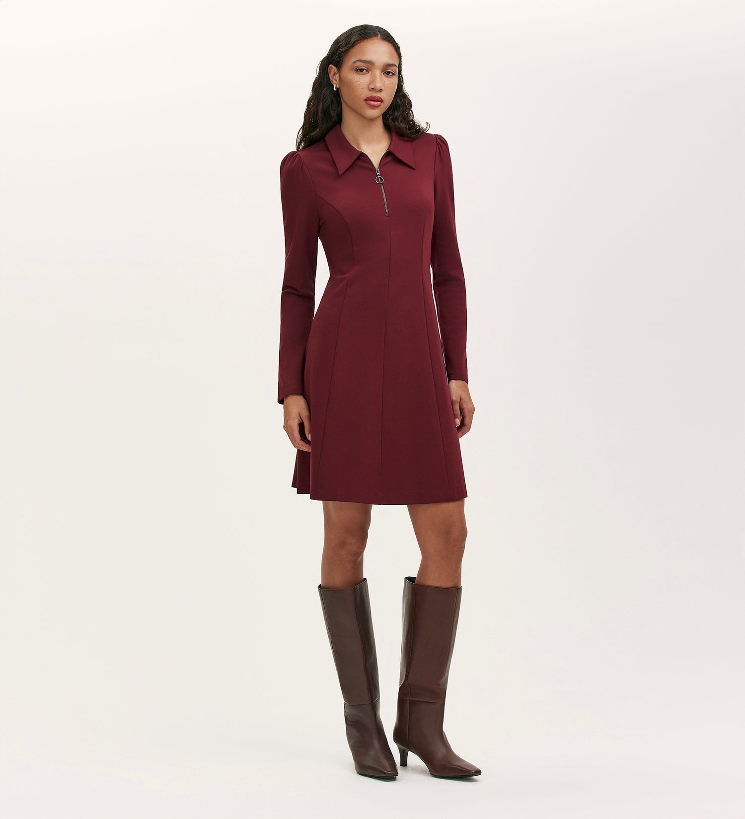 A woman stands against a plain light background wearing the Lea Burgundy Ponte Jersey Short Dress with a collared neckline and zipper, paired with tall dark brown heeled boots.