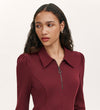 A woman with long, wavy brown hair and freckles wears the Lea Burgundy Ponte Jersey Short Dress with a collared neckline and zippered front, looking to the side against a plain light background.