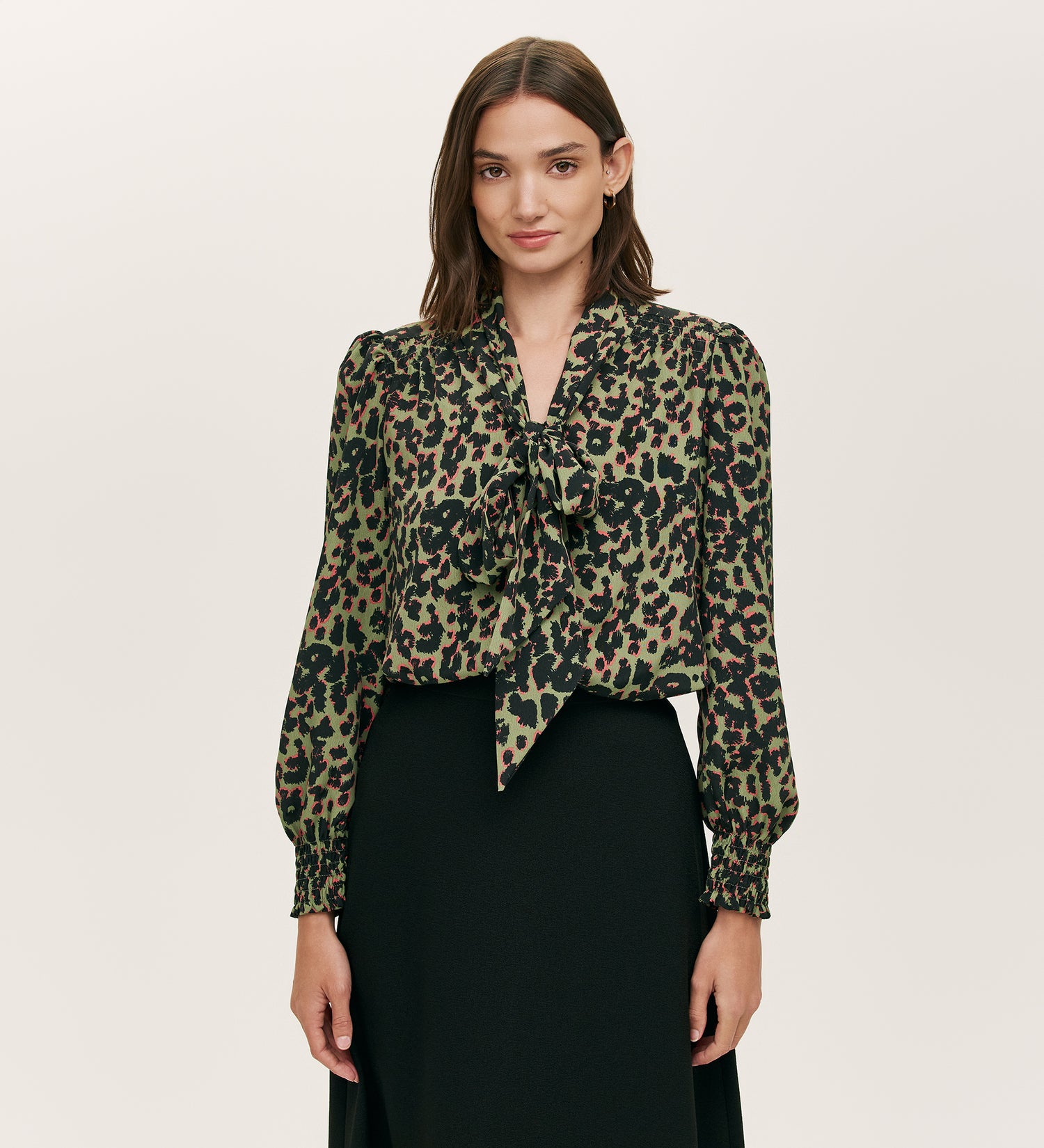A woman with straight brown hair wears the Lindsay Khaki Animal Print Pussy Bow Blouse with long, puffed sleeves and a black skirt, standing against a plain light background.