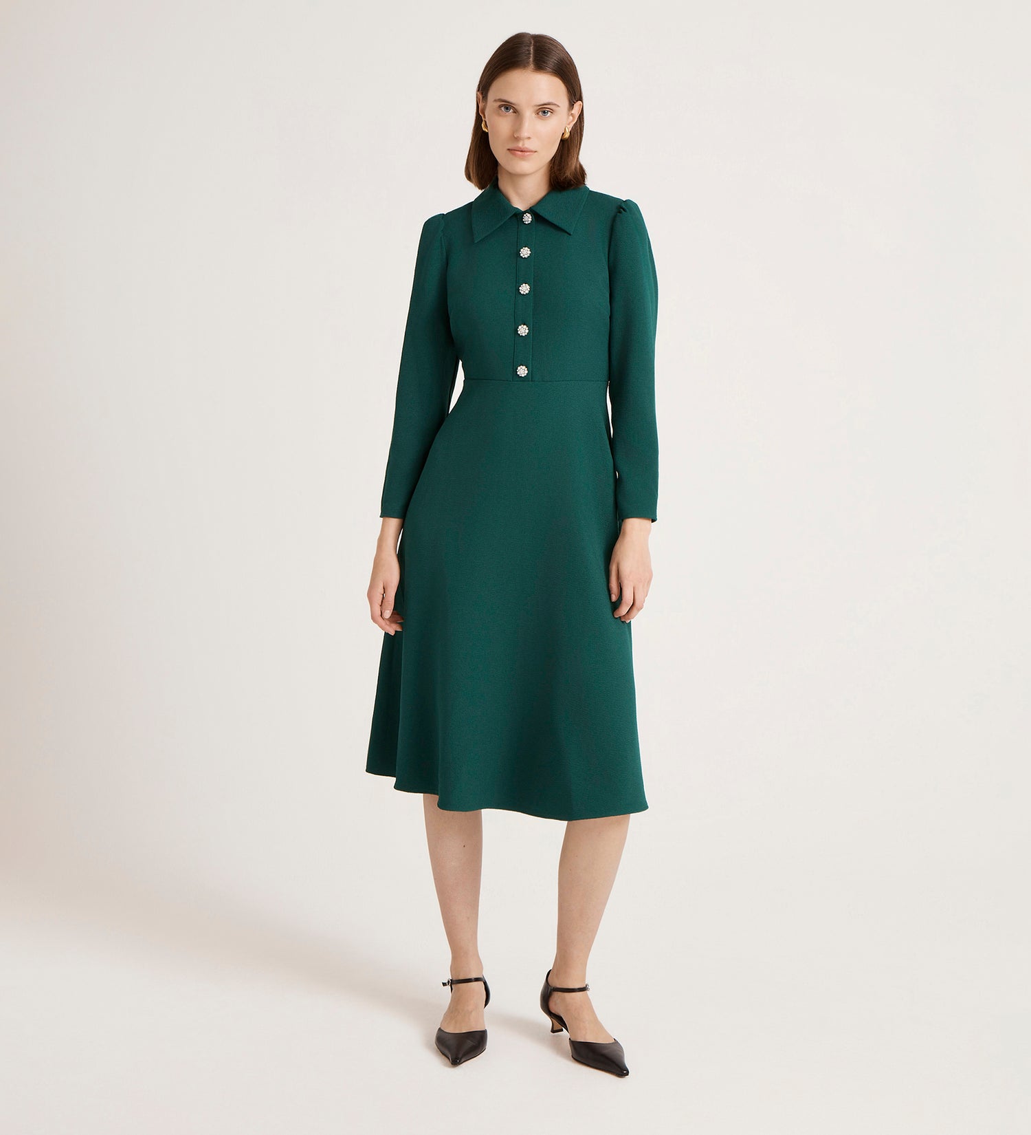A woman poses against a plain backdrop in the Lucia Bottle Green Crepe Midi Dress, featuring a fitted silhouette, long sleeves, classic collar, buttoned front, and paired with black ankle-strap heels.