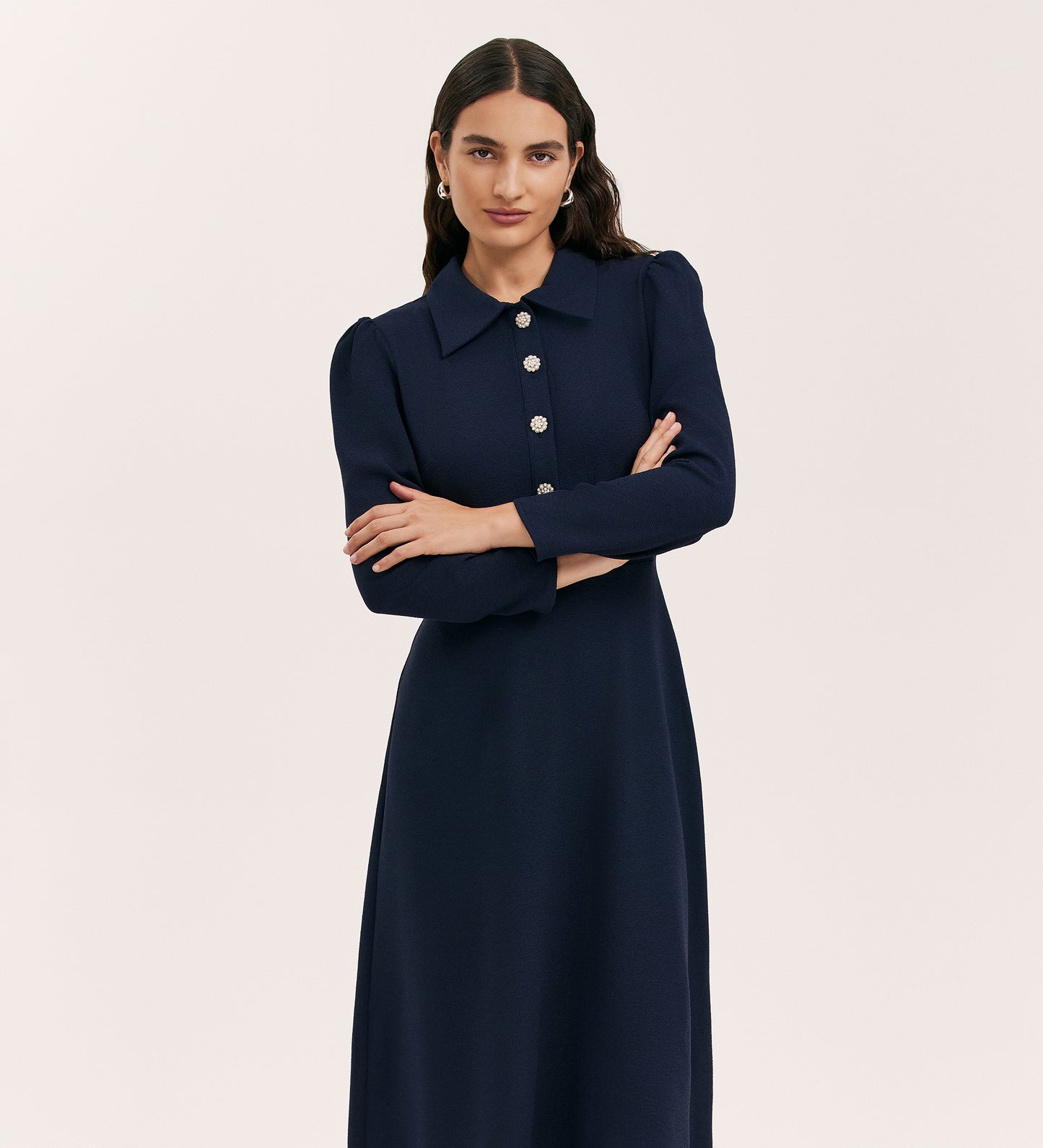 A woman with long dark hair stands with arms crossed against a plain light background, wearing the Lucia Navy Crepe Midi Dress—featuring crepe fabric, a classic collar, and decorative buttons.