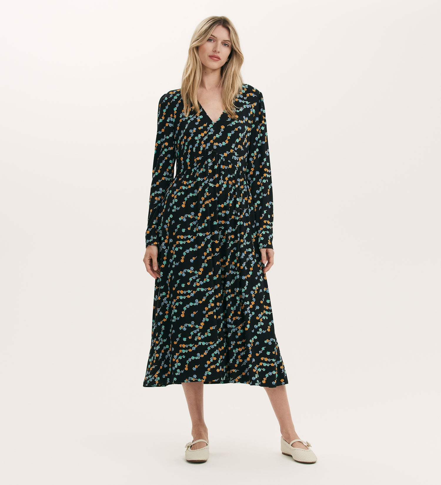 A woman stands against a plain light background wearing the Madelyn Black Rose Bud Midi Dress, featuring a V-neckline, long sleeves, black jersey fabric with blue, orange, and green floral print, paired with light-colored flats.