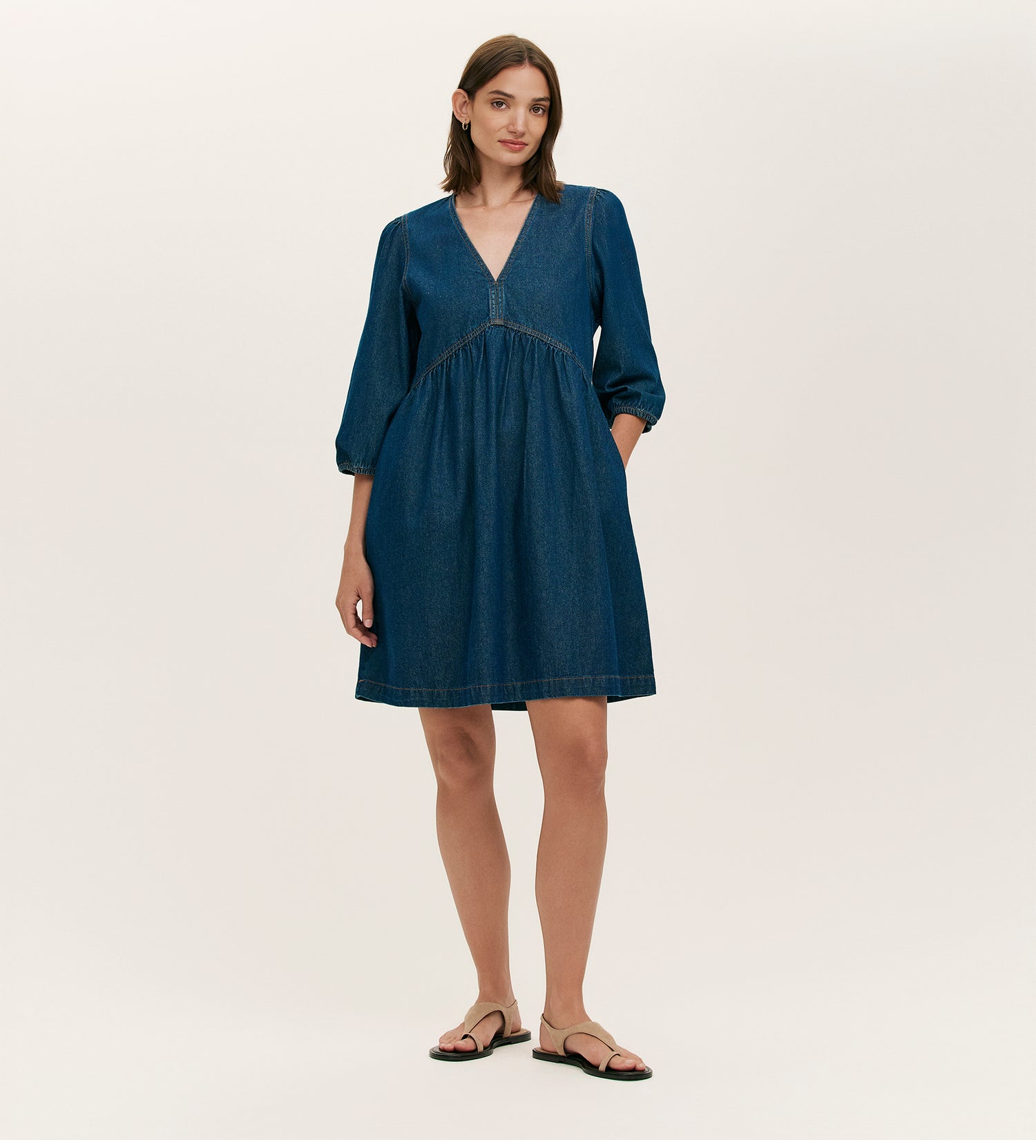 A woman with straight, shoulder-length brown hair and a relaxed expression stands against a plain background wearing the Marilyn Indigo Denim Short Dress with a V-neckline, three-quarter sleeves, and black sandals.