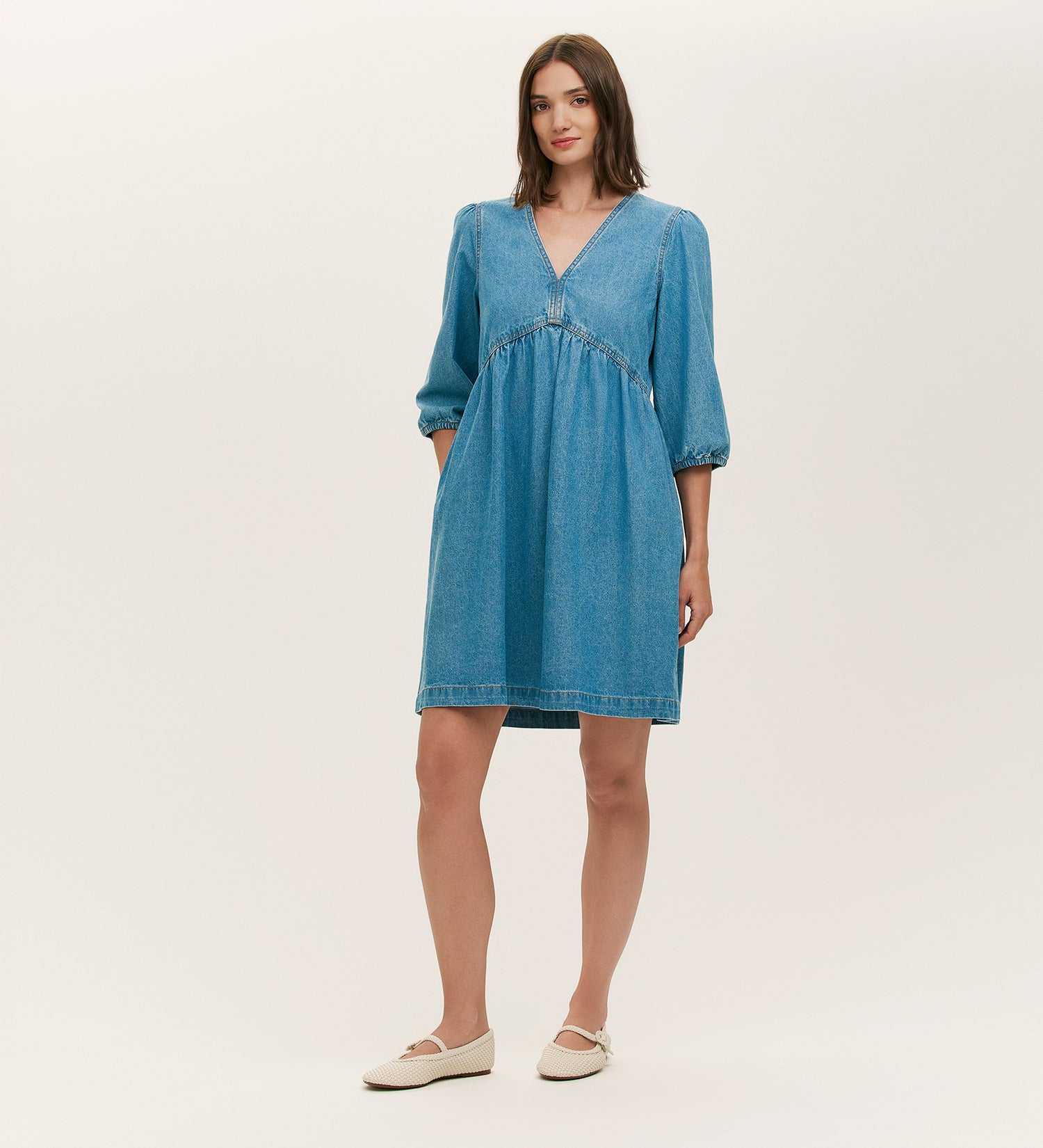 A woman wears the Marilyn Light Blue Denim Short Dress, featuring a relaxed fit, V-neckline, and three-quarter sleeves. The knee-length denim dress pairs with white flats. She has straight brown hair and stands against a plain light background.