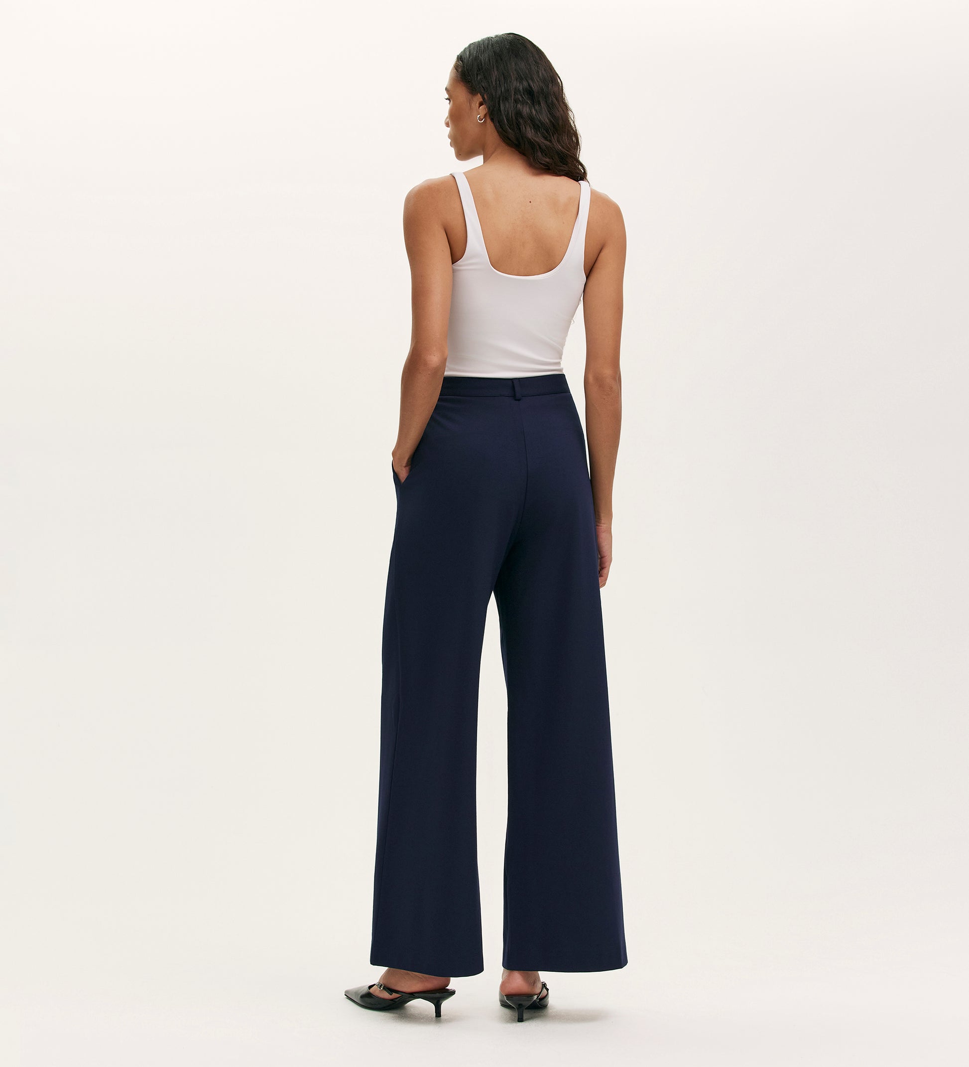 A woman with loose, dark, wavy hair stands facing away in a white tank top and Marissa Navy Wide Leg Trousers. Her hands are in her pockets, she wears black heels, and the background is plain and light.