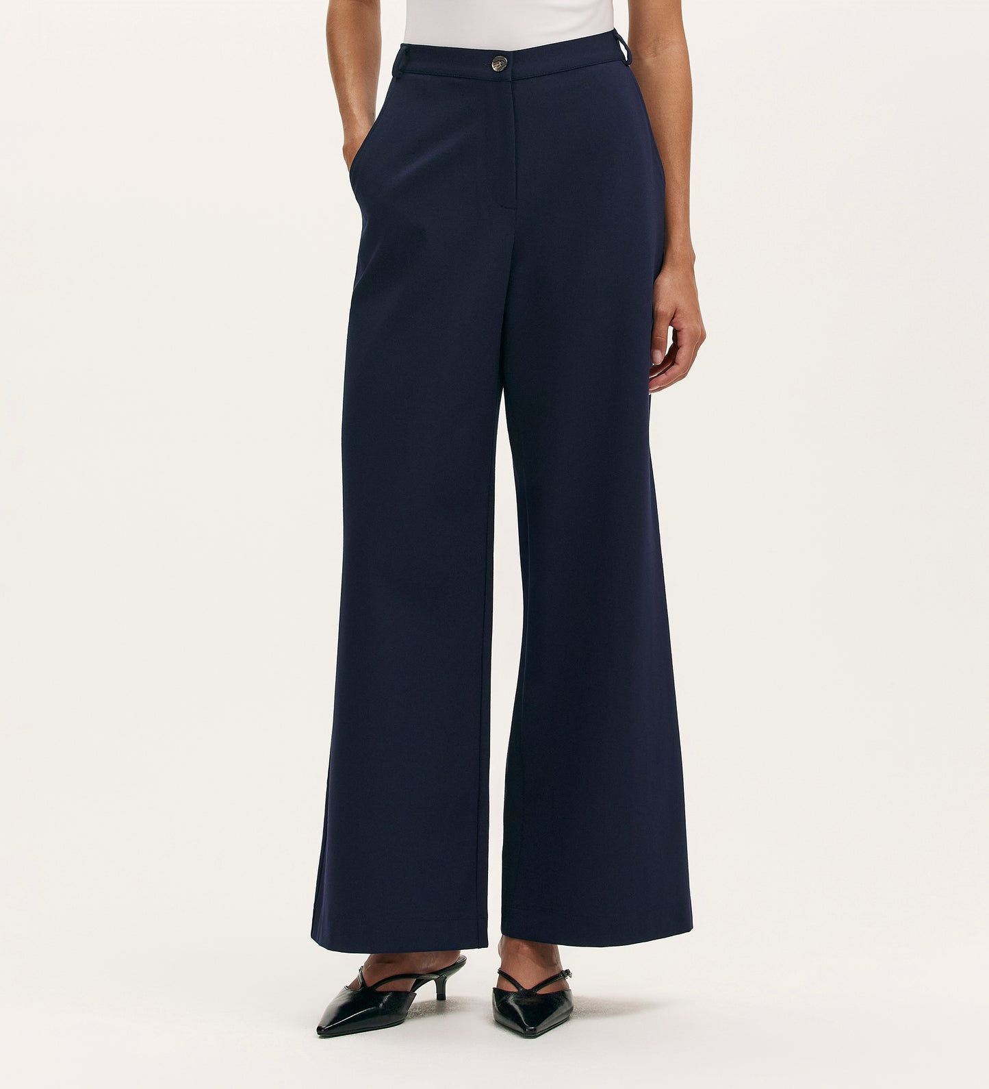 A person in Marissa Navy Wide Leg Trousers with a white top and black pointed-toe heels poses with hands in pockets against a plain light background.