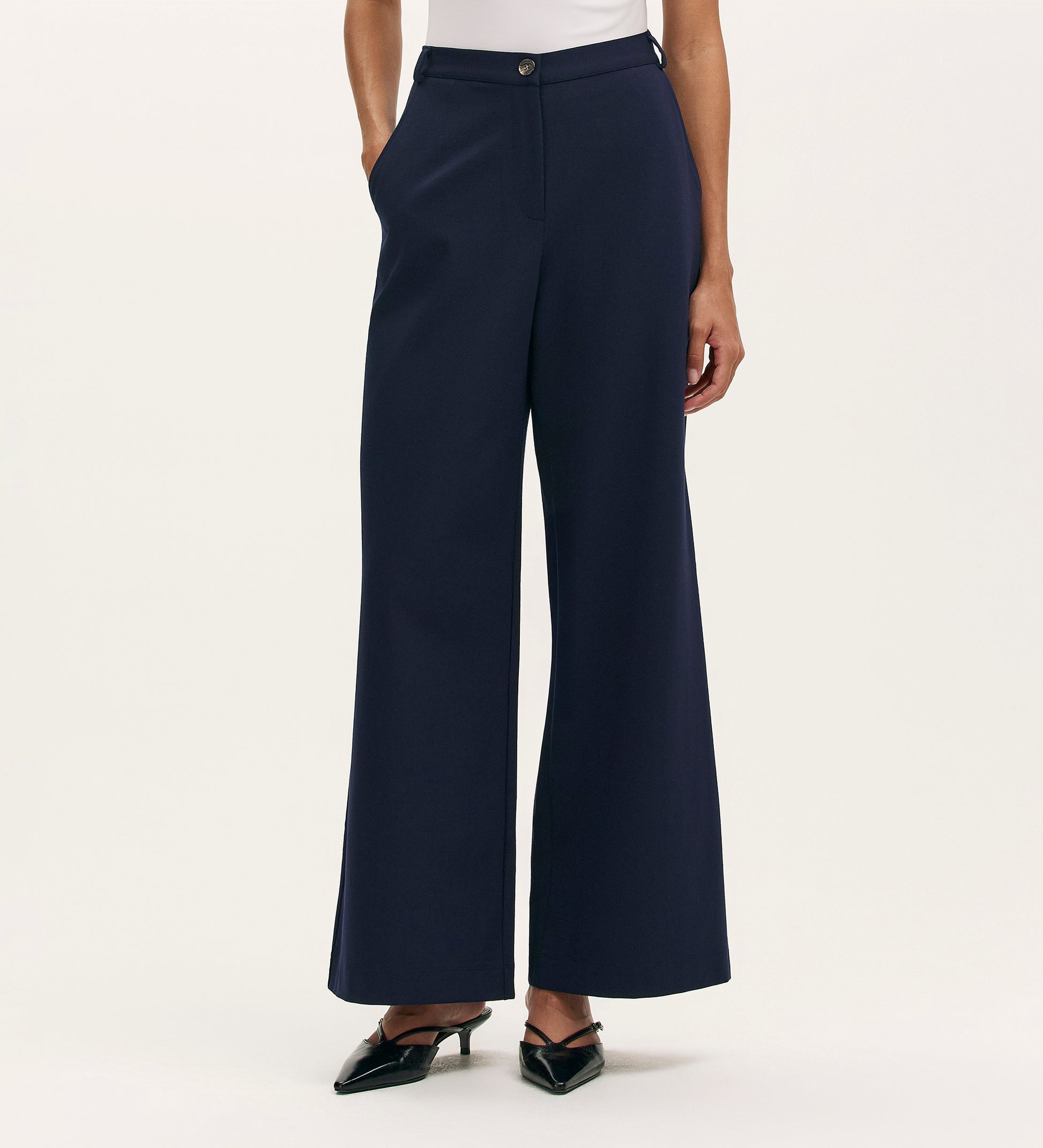 A person in Marissa Navy Wide Leg Trousers with a white top and black pointed-toe heels poses with hands in pockets against a plain light background.