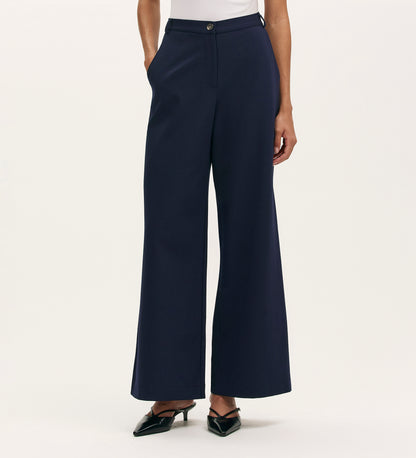A person in Marissa Navy Wide Leg Trousers with a white top and black pointed-toe heels poses with hands in pockets against a plain light background.