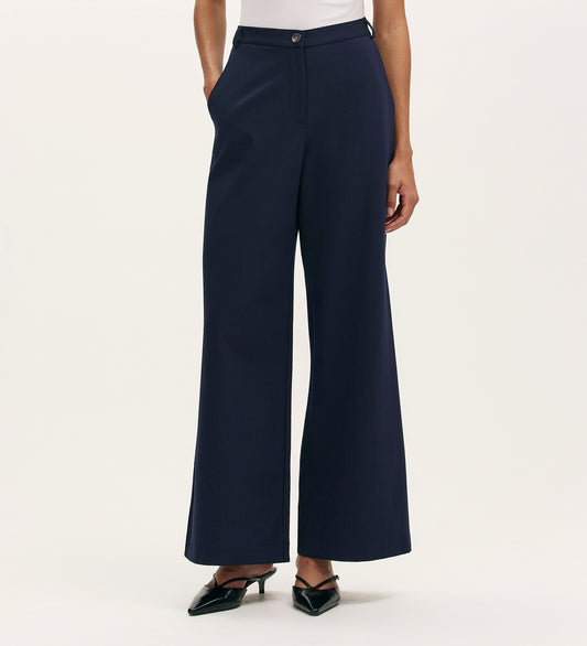 A person in Marissa Navy Wide Leg Trousers with a white top and black pointed-toe heels poses with hands in pockets against a plain light background.