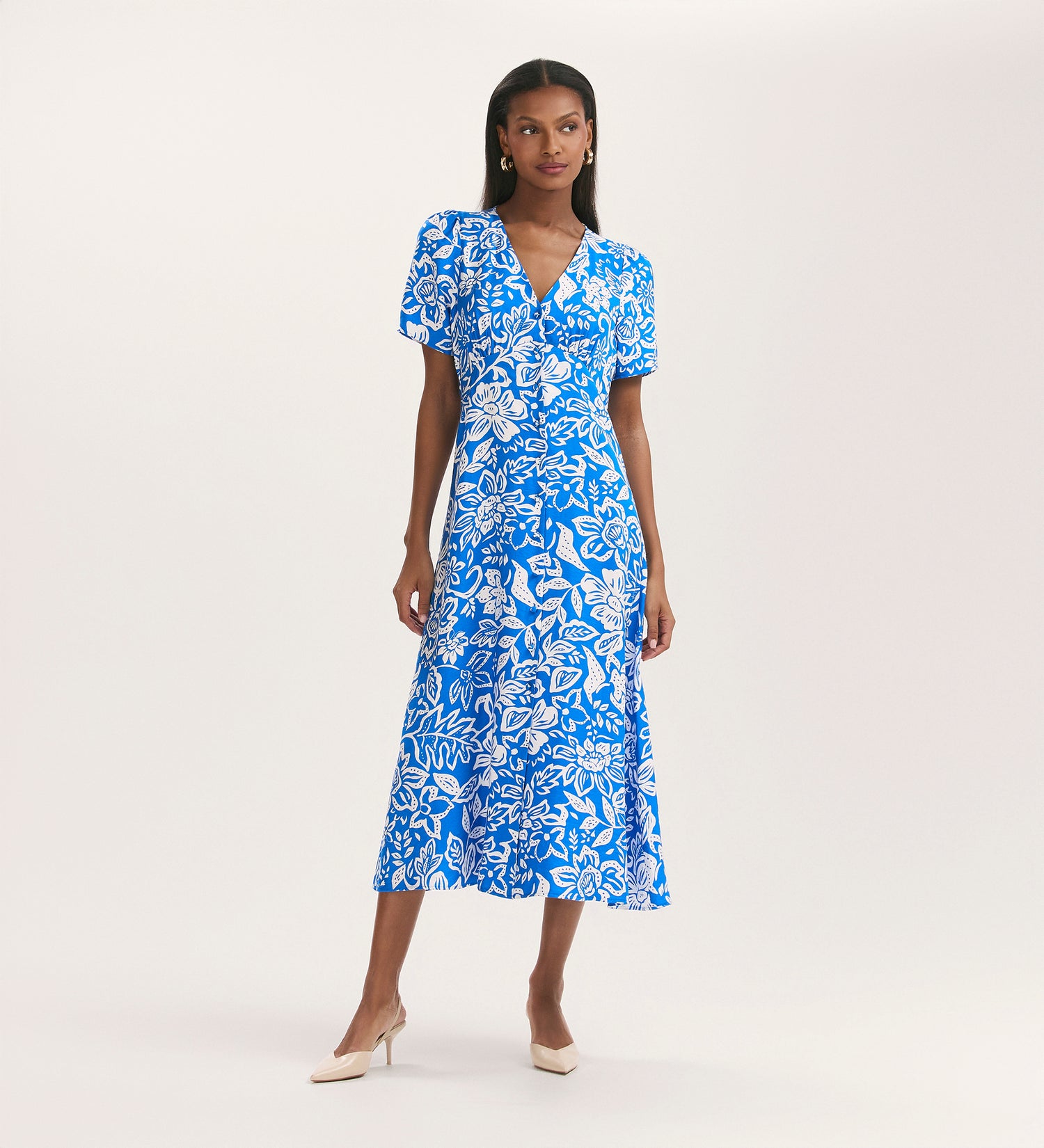 A woman poses against a plain light background wearing the Martina Blue Flower Crepe Midi Dress, featuring a button-down front, short sleeves, V-neck, and paired with beige heels.