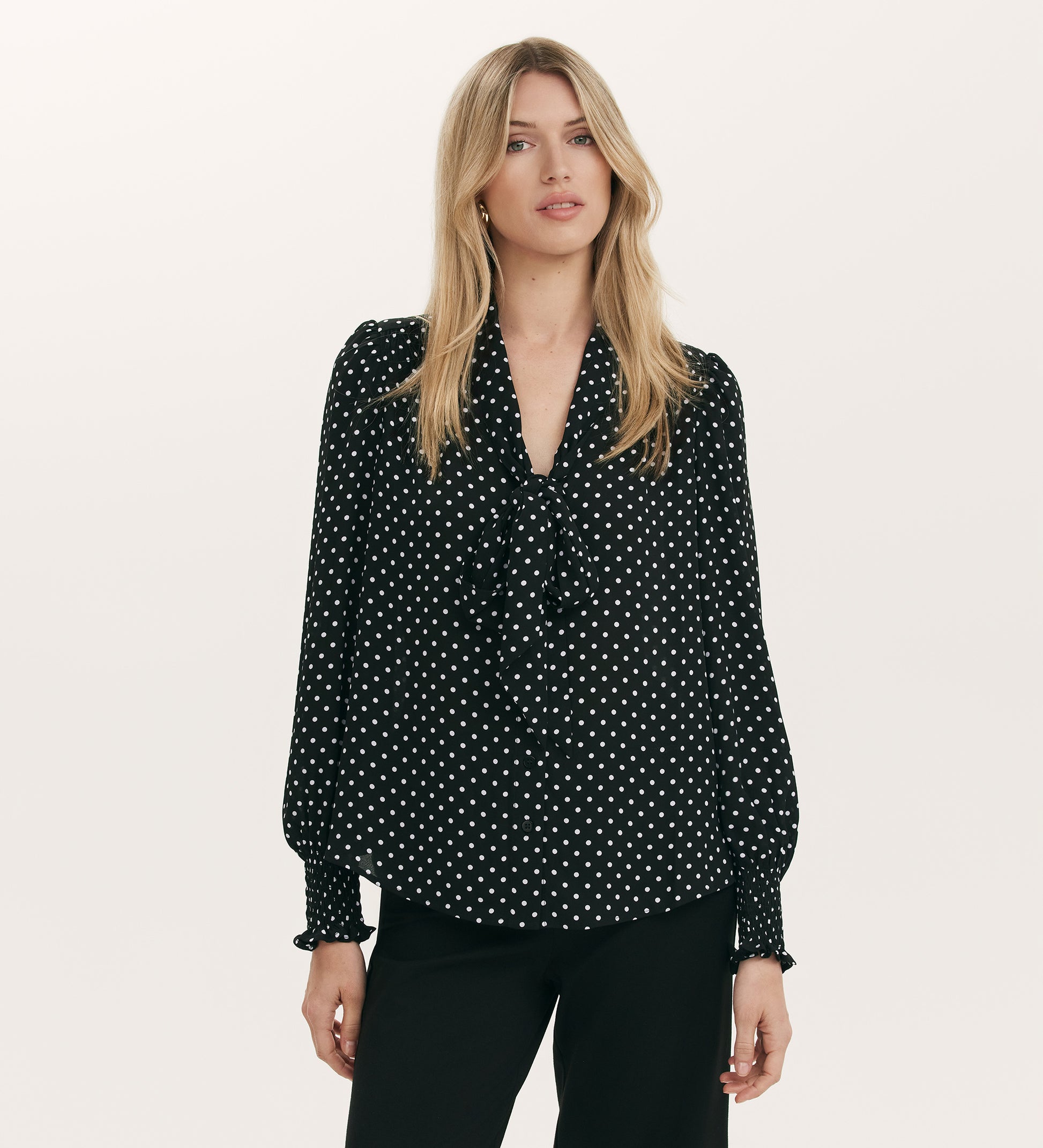 A woman with long blonde hair models the Mavis Black Polka Dot Crepe Tie Neck Blouse and black pants against a plain light background.