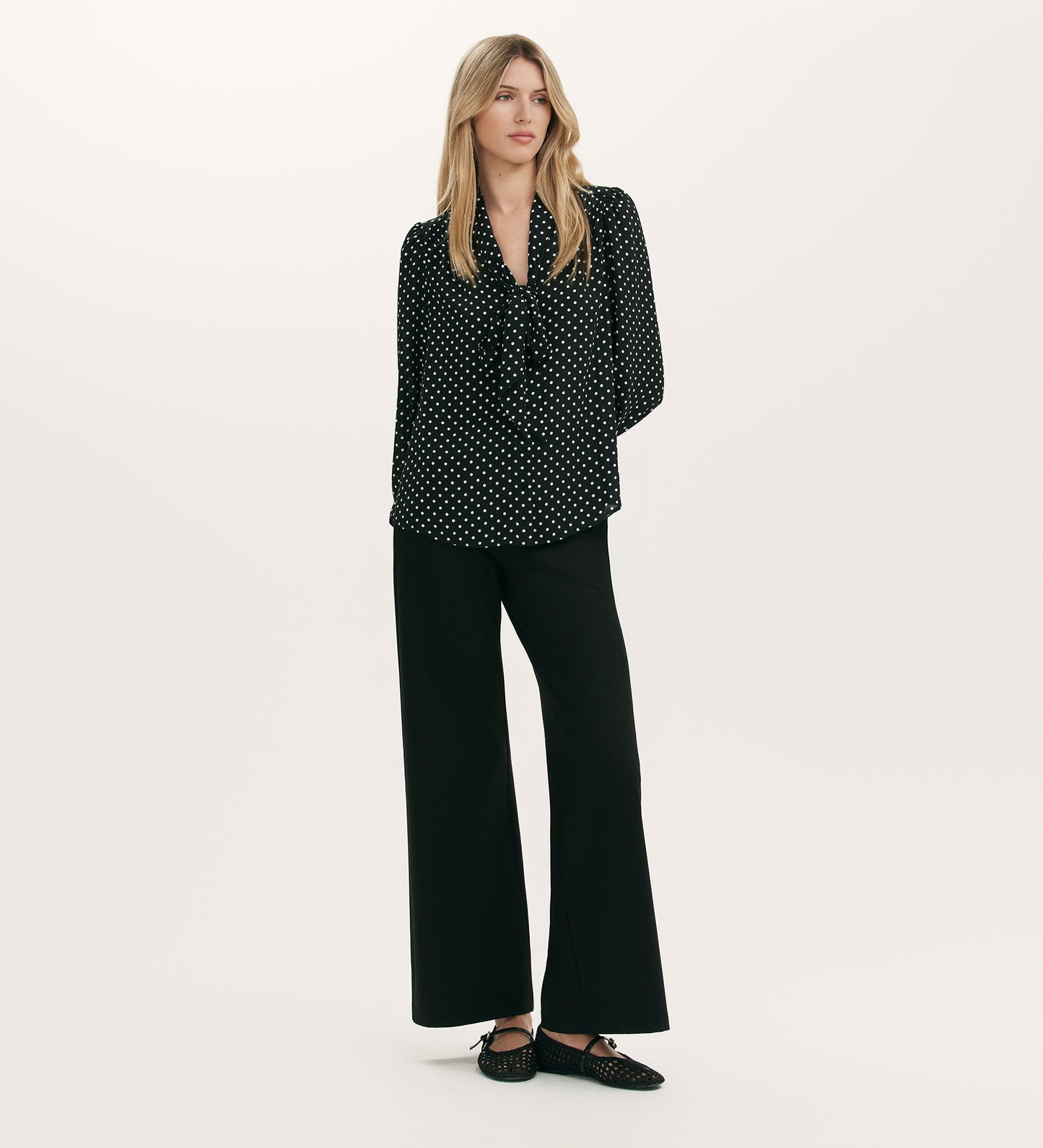 A woman with long blonde hair and a neutral expression stands against a plain background wearing the Mavis Black Polka Dot Crepe Tie Neck Blouse, black wide-leg pants, and black studded shoes.
