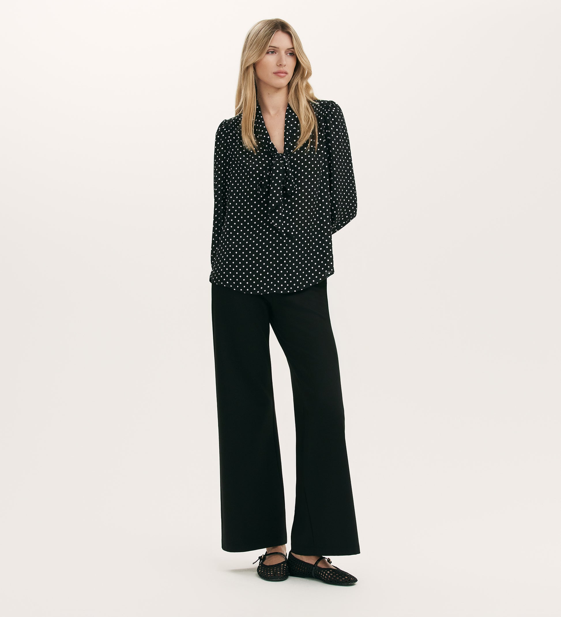A woman with long blonde hair and a neutral expression stands against a plain background wearing the Mavis Black Polka Dot Crepe Tie Neck Blouse, black wide-leg pants, and black studded shoes.