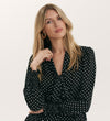 A woman with long blonde hair and gold hoop earrings models the Mavis Black Polka Dot Crepe Tie Neck Blouse, posing against a plain light background with one hand near her collar.