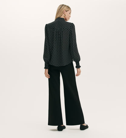 A woman with straight blonde hair stands facing away against a plain light backdrop, wearing the Mavis Black Polka Dot Crepe Tie Neck Blouse with long sleeves, paired with loose black pants and black shoes.