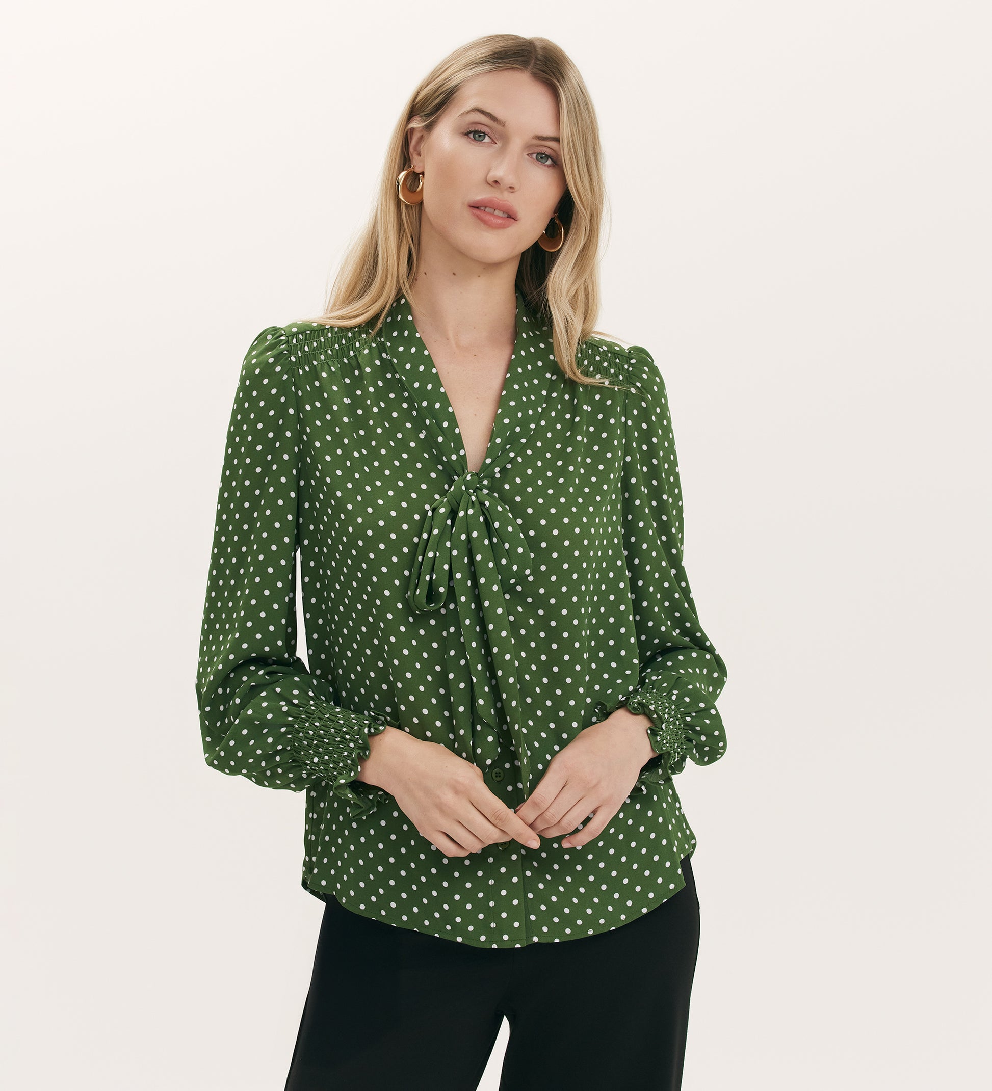 A woman with long blonde hair wears the Mavis Green Spot Chiffon Pussy Bow Blouse, featuring shirred cuffs and puffed sleeves, paired with black pants, standing against a plain light background.