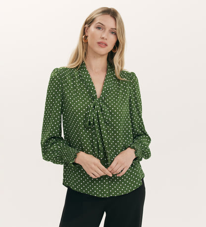 A woman with long blonde hair wears the Mavis Green Spot Chiffon Pussy Bow Blouse, featuring shirred cuffs and puffed sleeves, paired with black pants, standing against a plain light background.