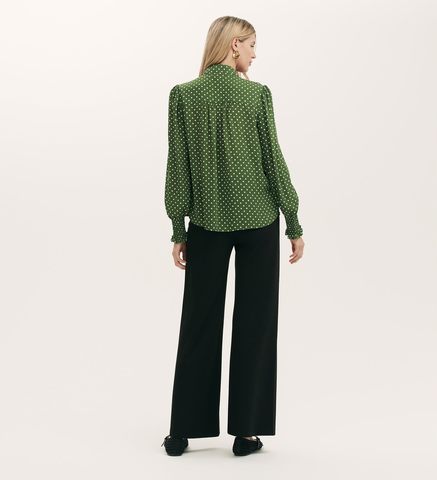 A woman with long blonde hair, seen from behind, wears the Mavis Green Spot Chiffon Pussy Bow Blouse and black wide-leg pants with black shoes, set against a plain light background.