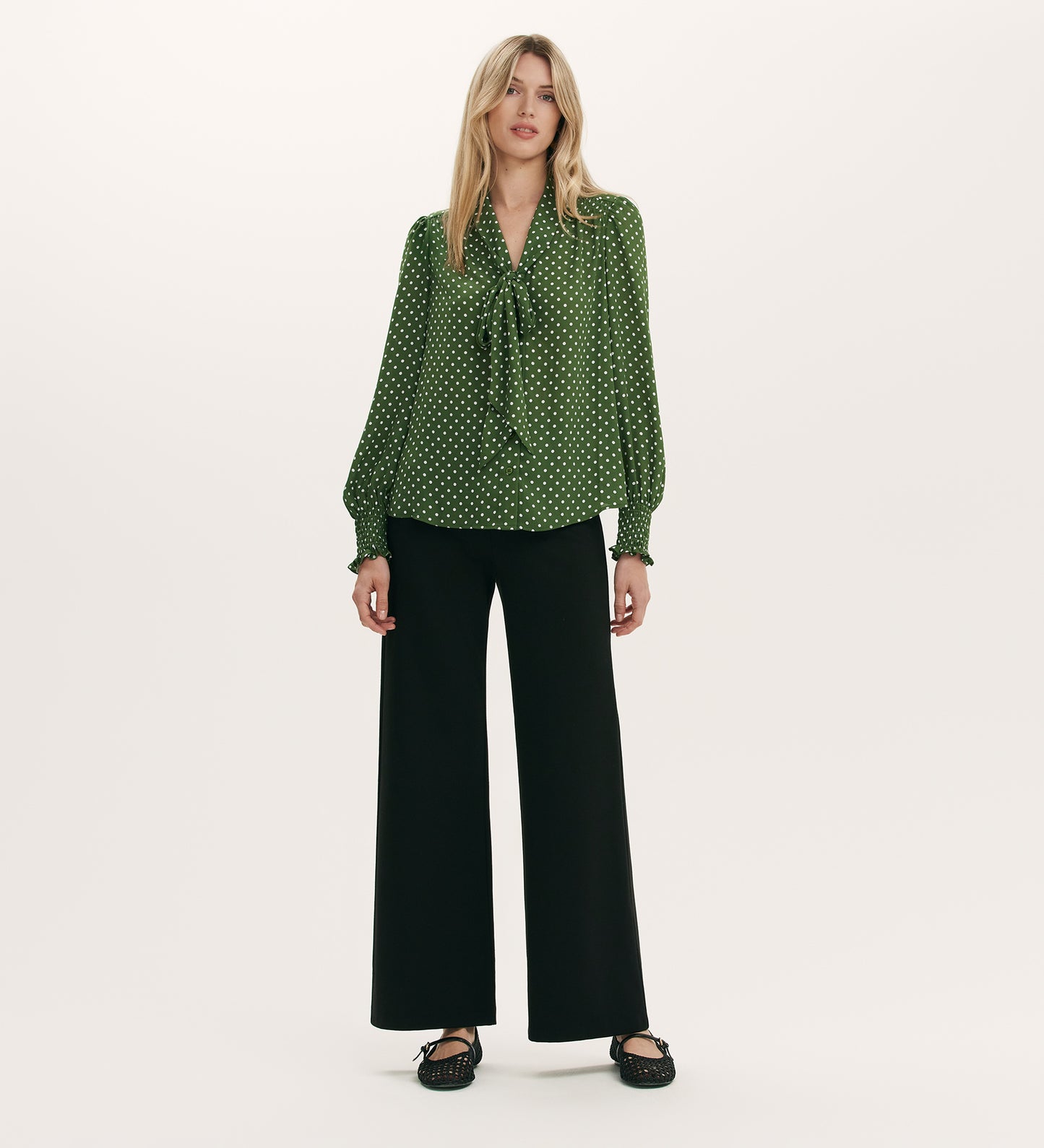 A woman stands against a plain background wearing the Mavis Green Spot Chiffon Pussy Bow Blouse with black wide-leg pants and black shoes.