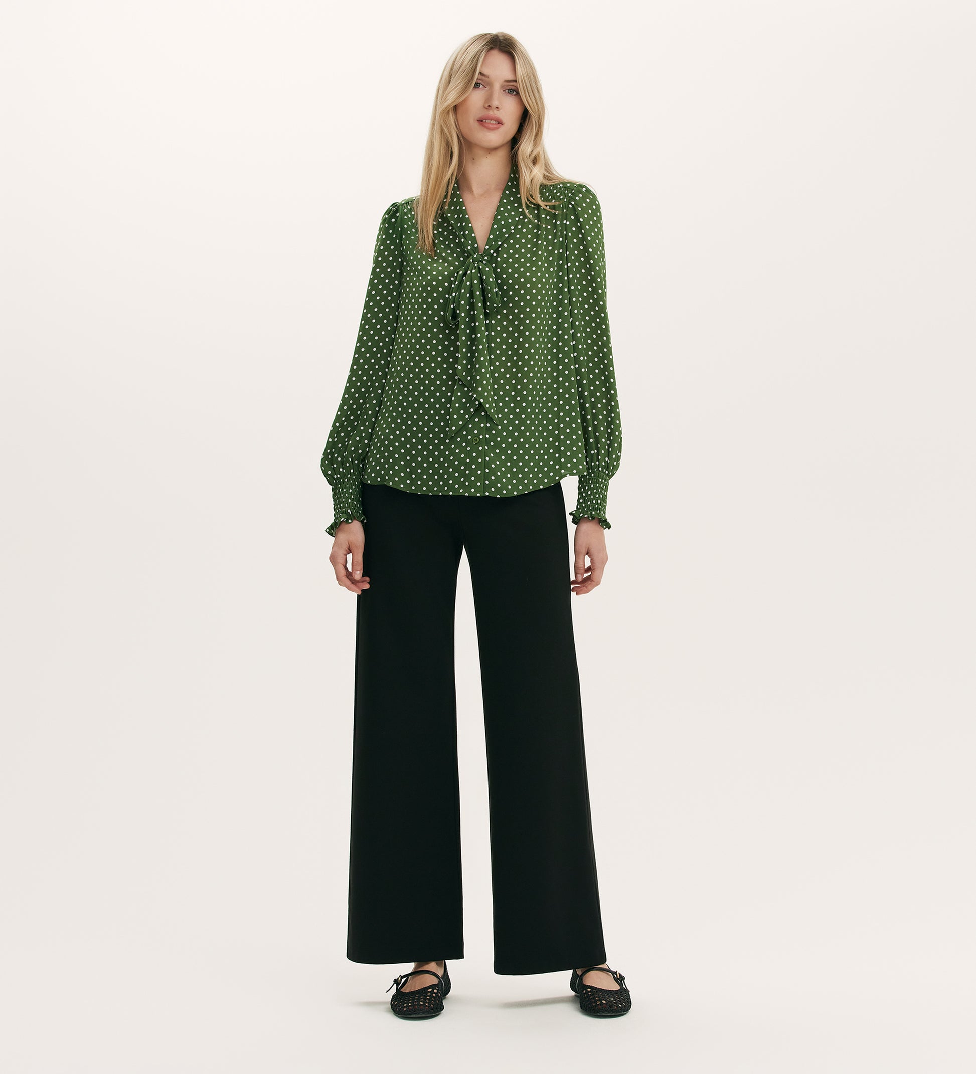 A woman stands against a plain background wearing the Mavis Green Spot Chiffon Pussy Bow Blouse with black wide-leg pants and black shoes.