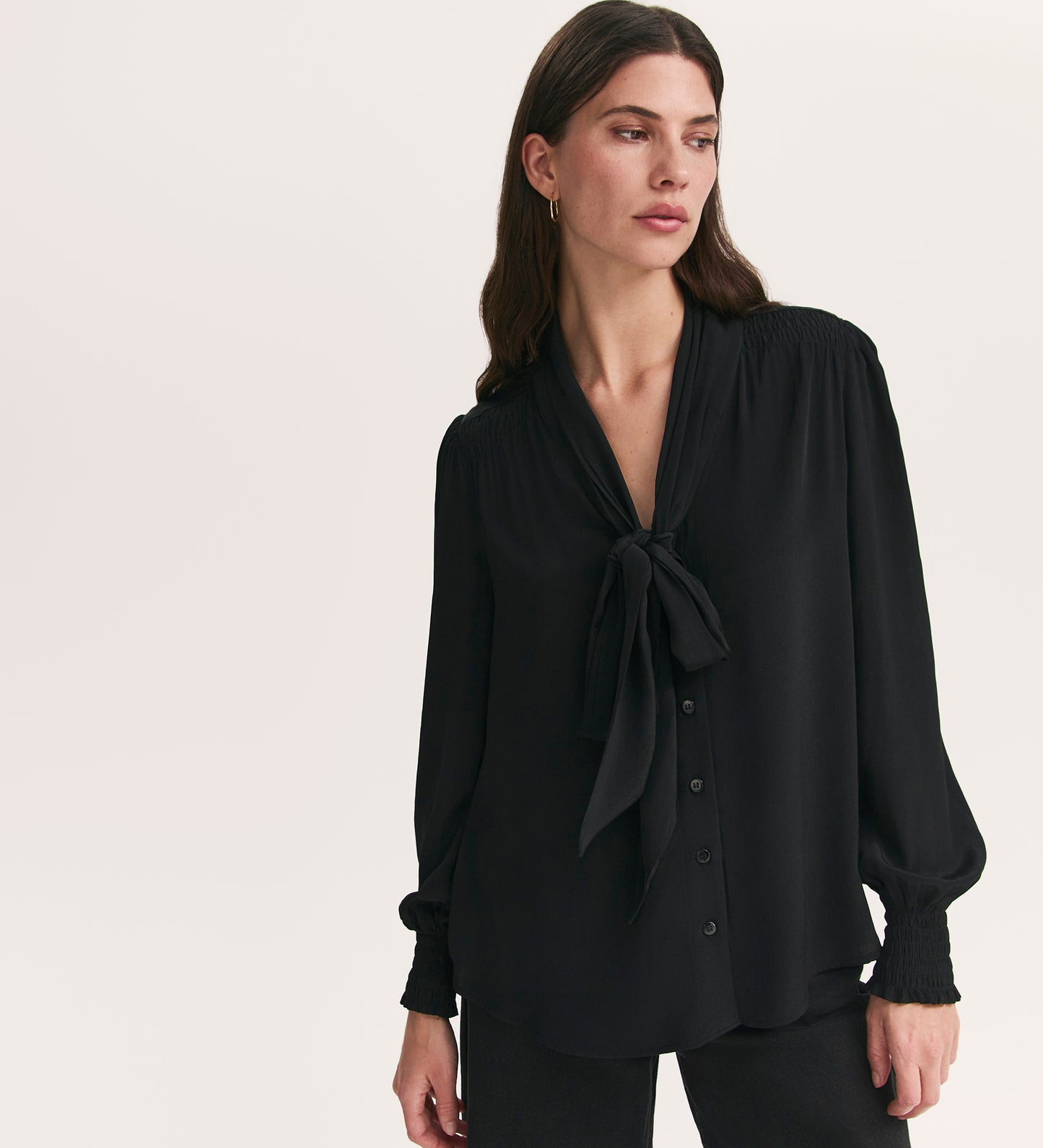 A woman with long brown hair wears the elegant Mavis Black Chiffon Blouse, a loose-fitting pussy bow top, paired with black pants against a plain light background.